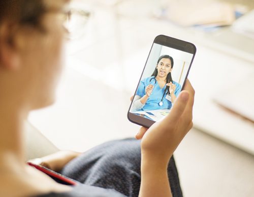 Photo of a telemedicine session. A woman sits on a couch holding a cell phone . On the cell phone screen is a female medical provider with long dark hair wearing blue scrubs and a stethoscope. Her mouth and hands are in motion as if she is speaking to the woman holding the phone.