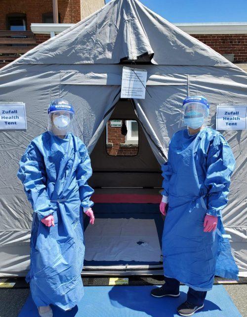 Photo of two medical personnel dressed in personal protective equipment standing in front of a medical tent