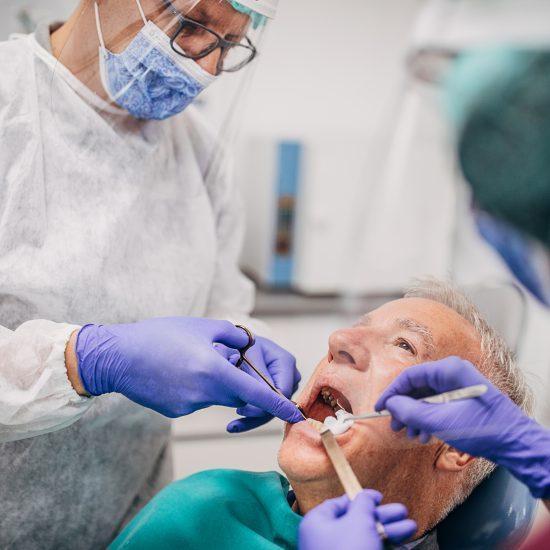 Male dentist and his female assistant wearing a protective gowns, eyewear protection, protective face masks and gloves while working on the teeth of an elderly male patient