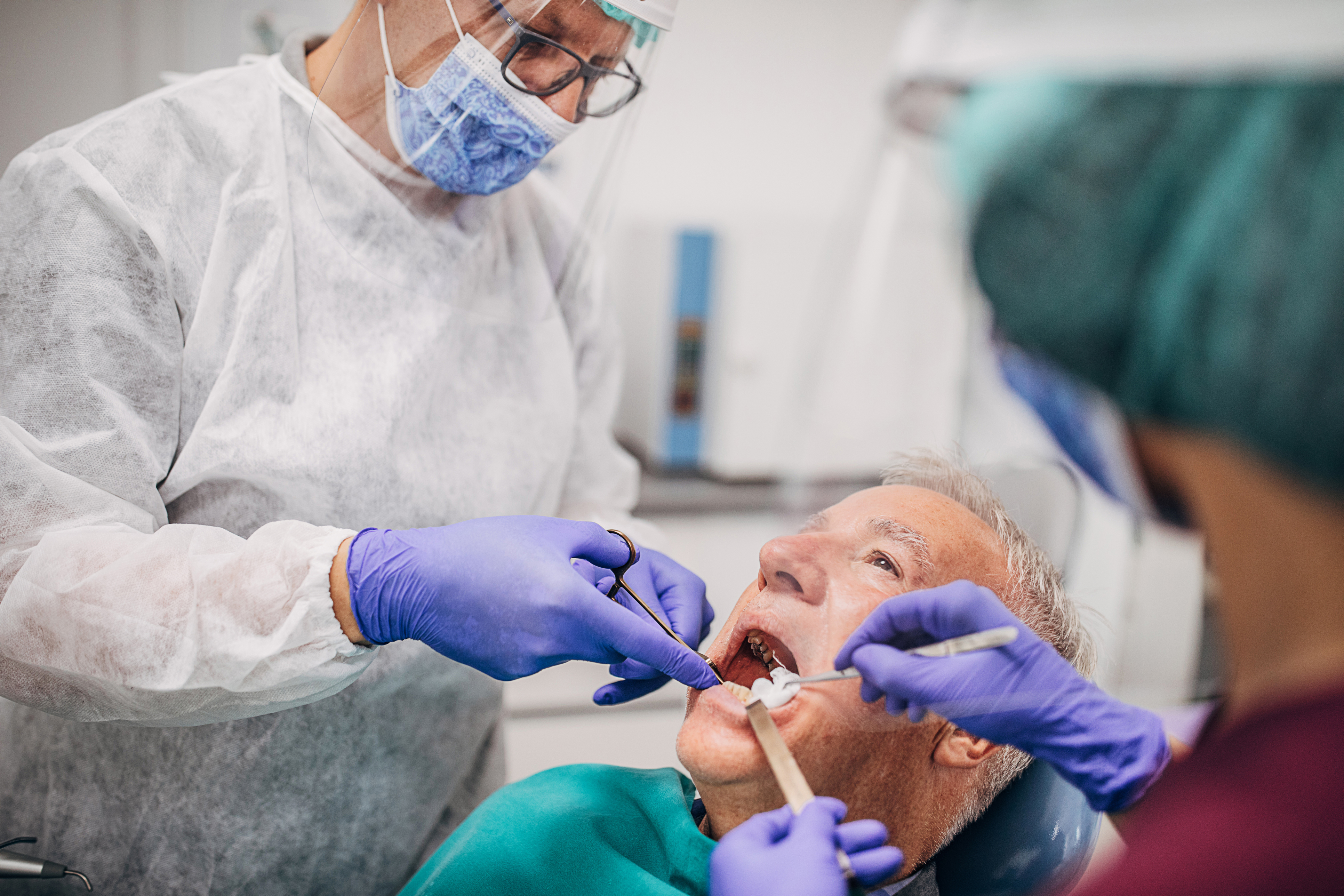 Male dentist and his female assistant wearing a protective gowns, eyewear protection, protective face masks and gloves while working on the teeth of an elderly male patient