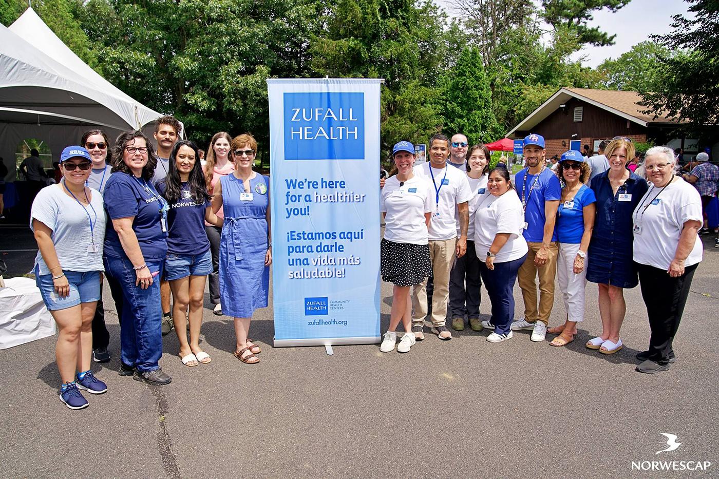 A large group of people stand around a banner that says "Zufall Health: We're here for a healthier you!" in a parking lot outside.