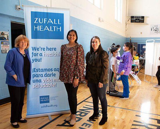 Three people, including Eva Turbiner on far left, stand smiling in front of a roll-up banner in a gymnasium. In the background, children receive medical care.