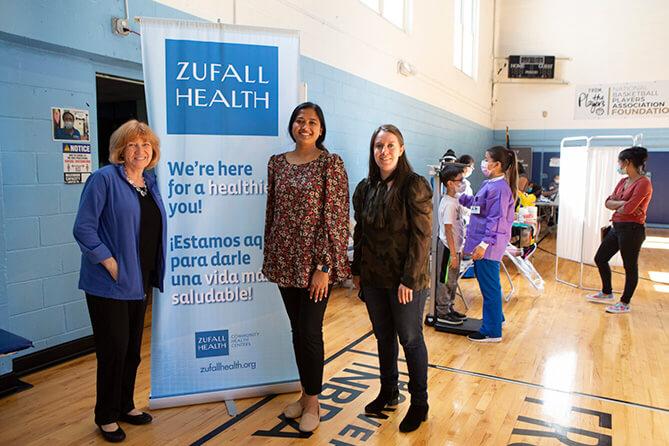 Three people, including Eva Turbiner on far left, stand smiling in front of a roll-up banner in a gymnasium. In the background, children receive medical care.