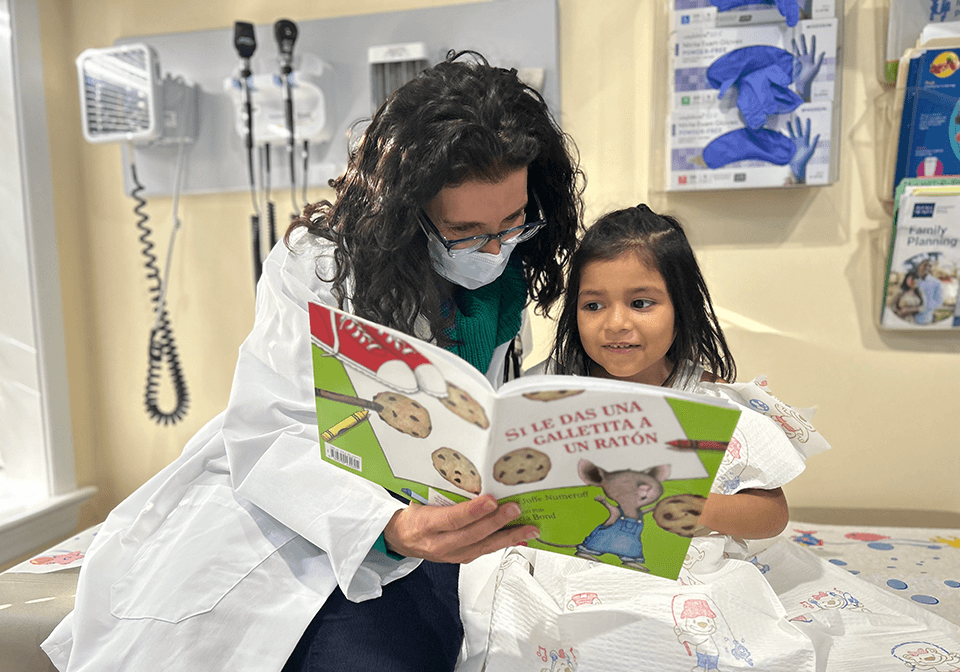 Doctor wearing a face masks reads a book with a young patient on an exam table during a well visit